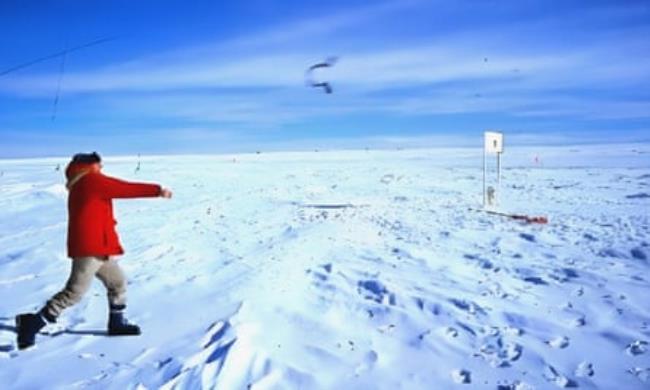 A man wearing a red coat throws a boomerang against a background of snowy ground and blue sky